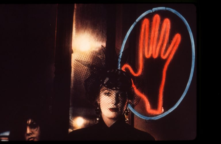 Woman in hat with veil standing in front of neon sign depicting a hand