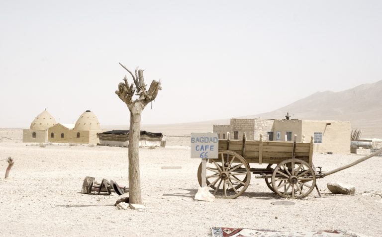 Desert scene with building in background and cart in foreground next to sign reading 
