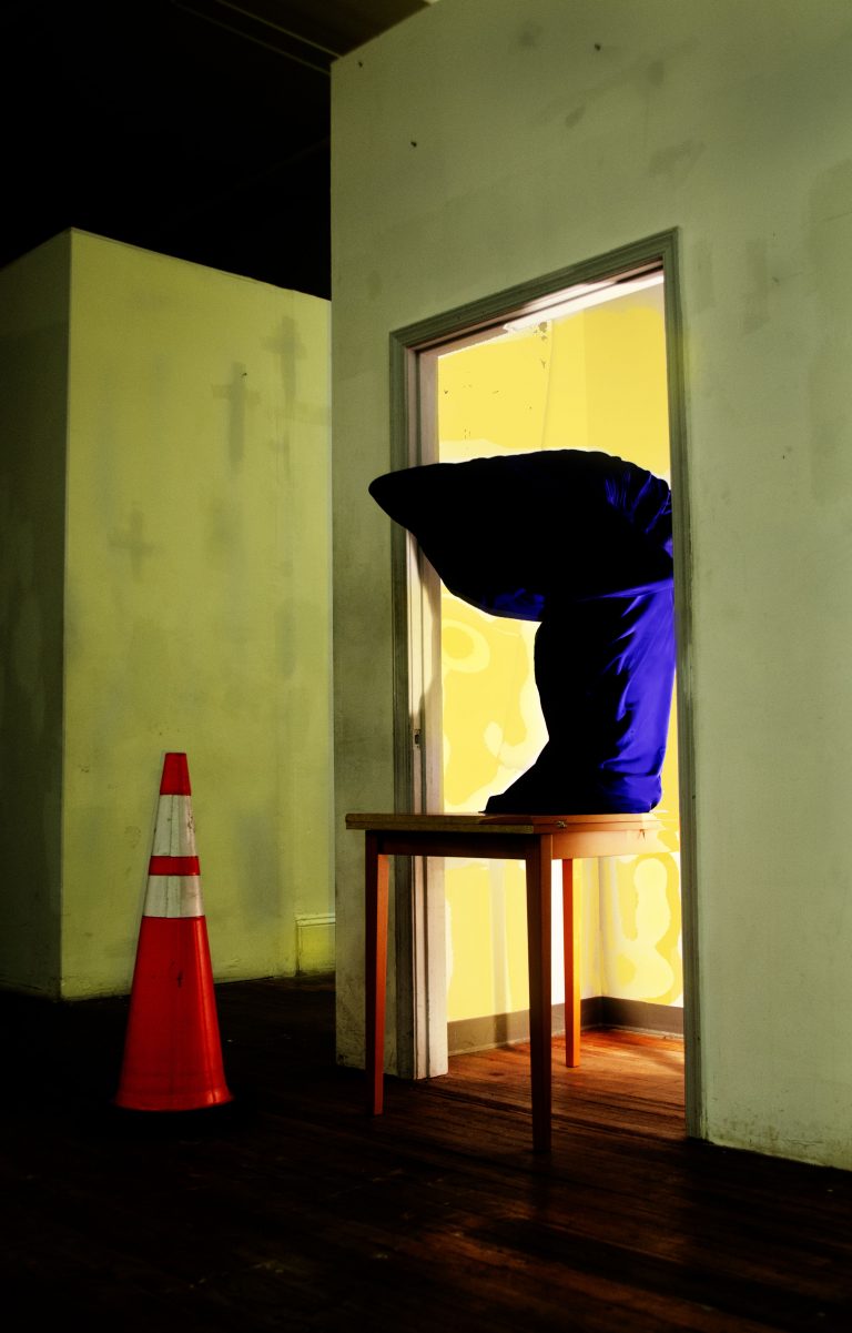Slouched figure wrapped in purple on table in illuminated doorway with traffic cone