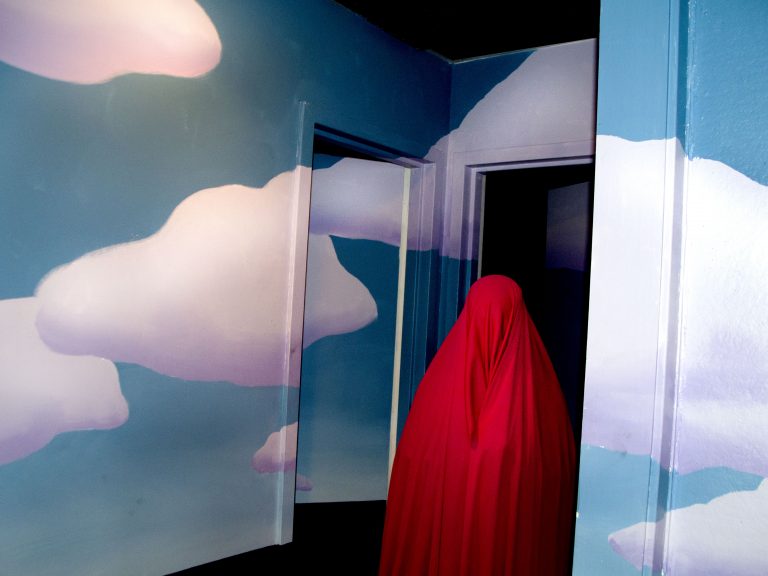 Figure shrouded in dark pink fabric in doorway painted with sky and clouds