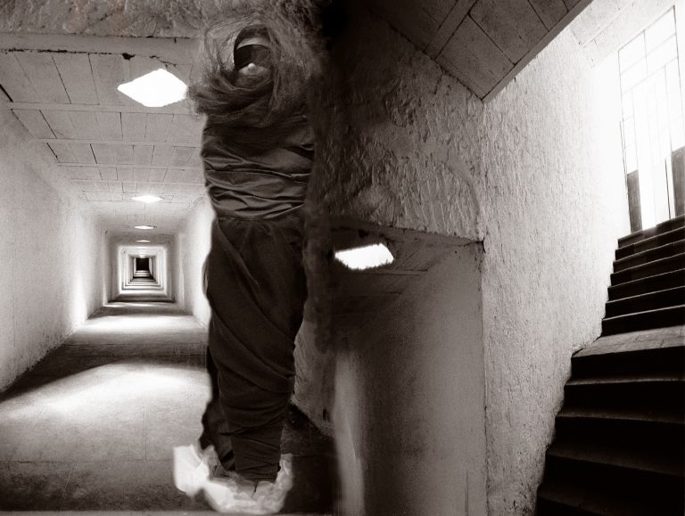 Black and white image of shrouded figure at bottom of stairs in a long hallway