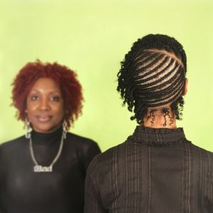 two black women, one faces away from the camera to show an intricate hairstyle, the other faces the camera in the background, in soft focus