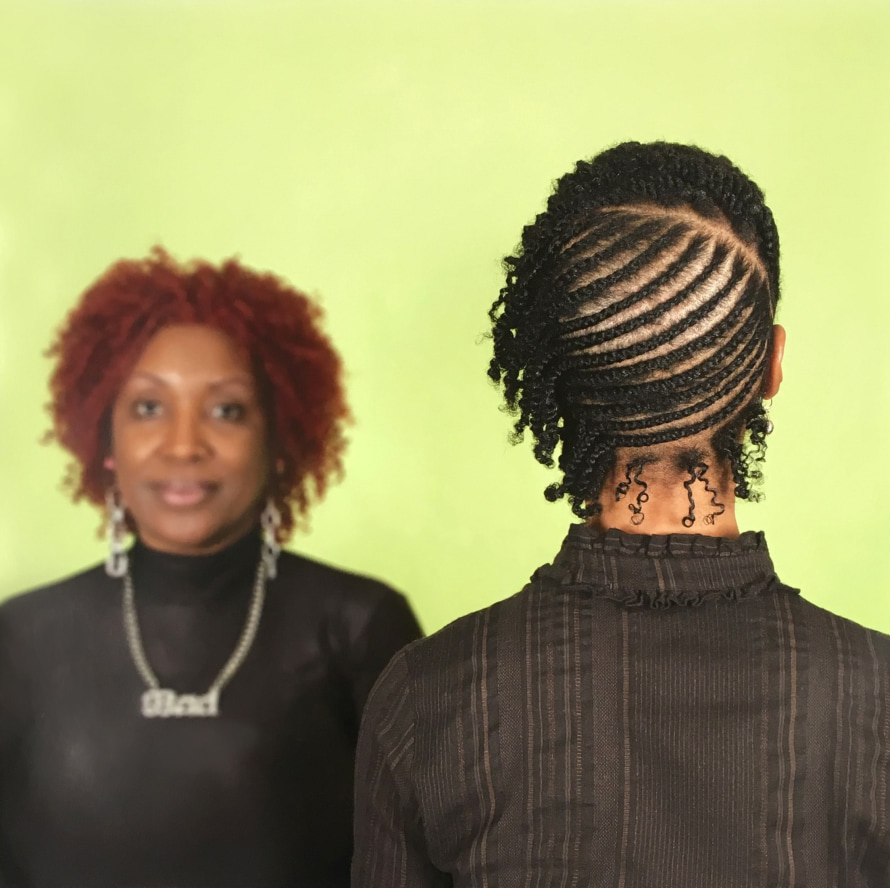 two black women, one faces away from the camera to show an intricate hairstyle, the other faces the camera in the background, in soft focus