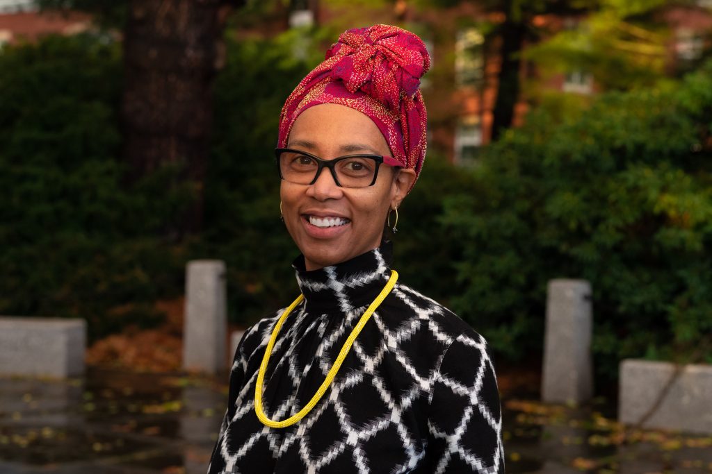 A Black woman is standing outside and smiling at the camera. She is wearing a red wrap on her head and a black and white top