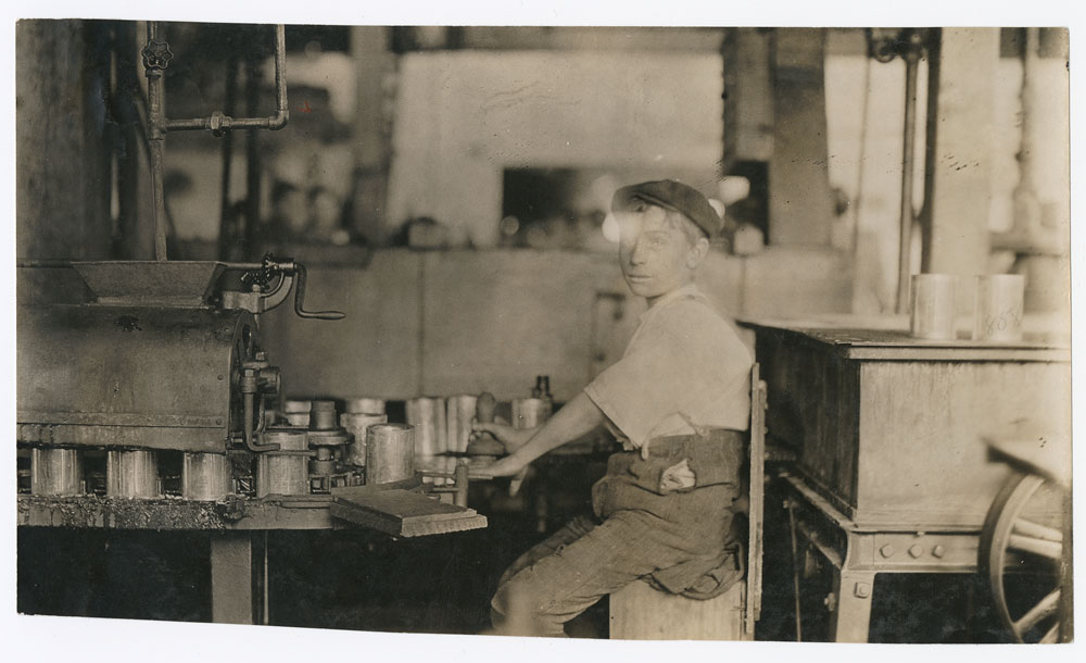 Sepia photograph of a boy sitting at a canning machine.
